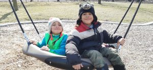 Twins, Alexandria and Sebastian are on a playground sitting side-by-side on a large swing and smiling.